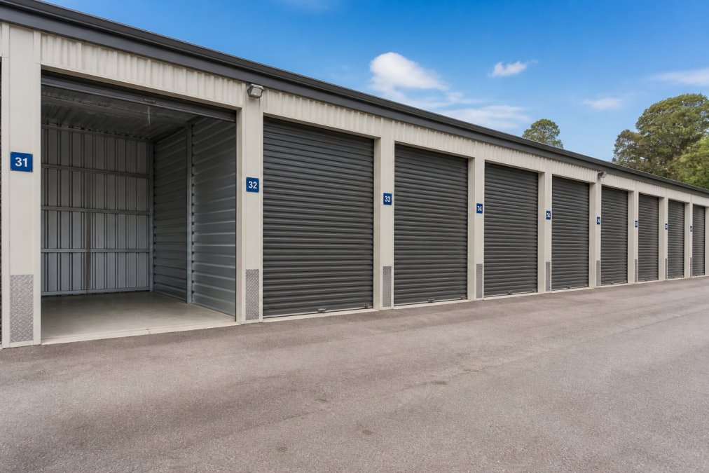 Interior view of a clean, well-lit self storage facility showing multiple unit doors of varying sizes arranged in a row, with drive-up access visible and numbered units clearly marked