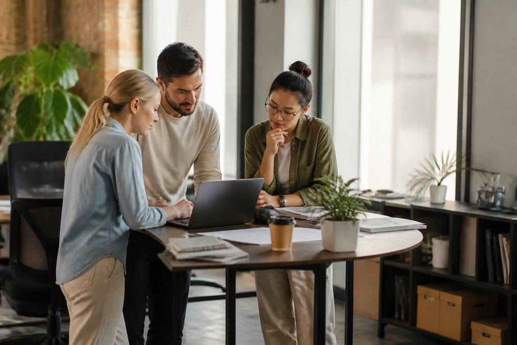 Small marketing team collaborating at a standing desk, reviewing SEO metrics and strategy on multiple monitors, modern office with natural lighting