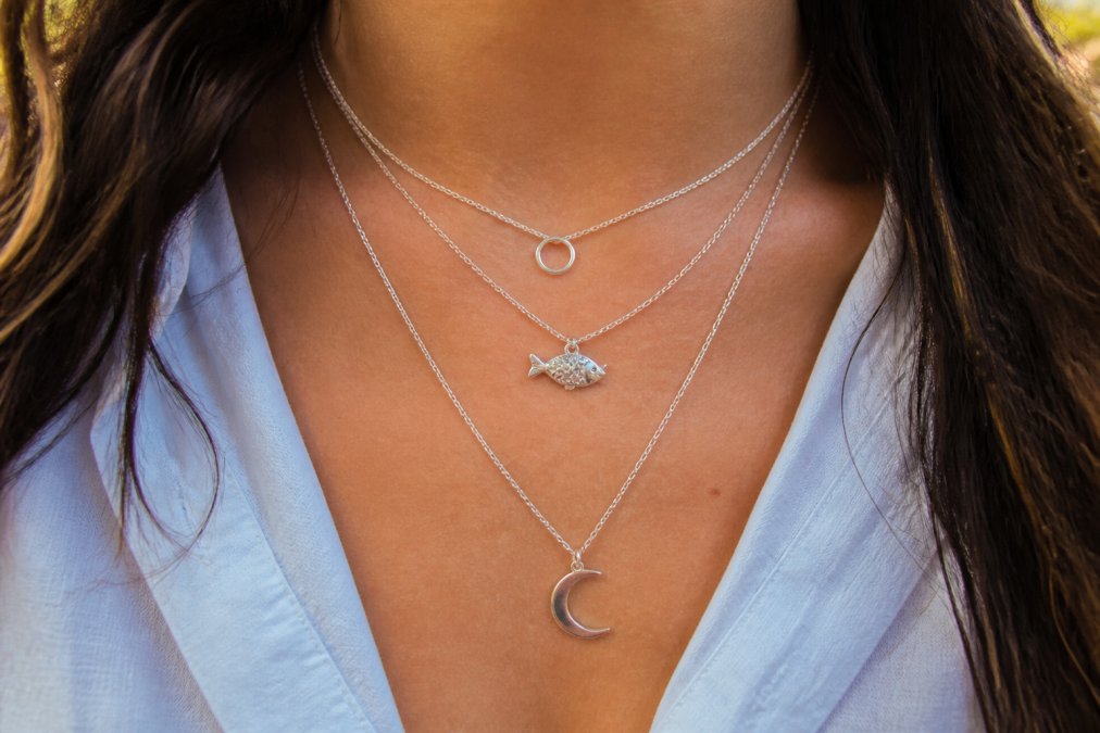 Close-up of a person wearing three delicate silver necklaces of varying lengths layered against their neck and chest, showing how the Open Circle, Fish, and Lunar necklaces sit together at different depths, catching light differently