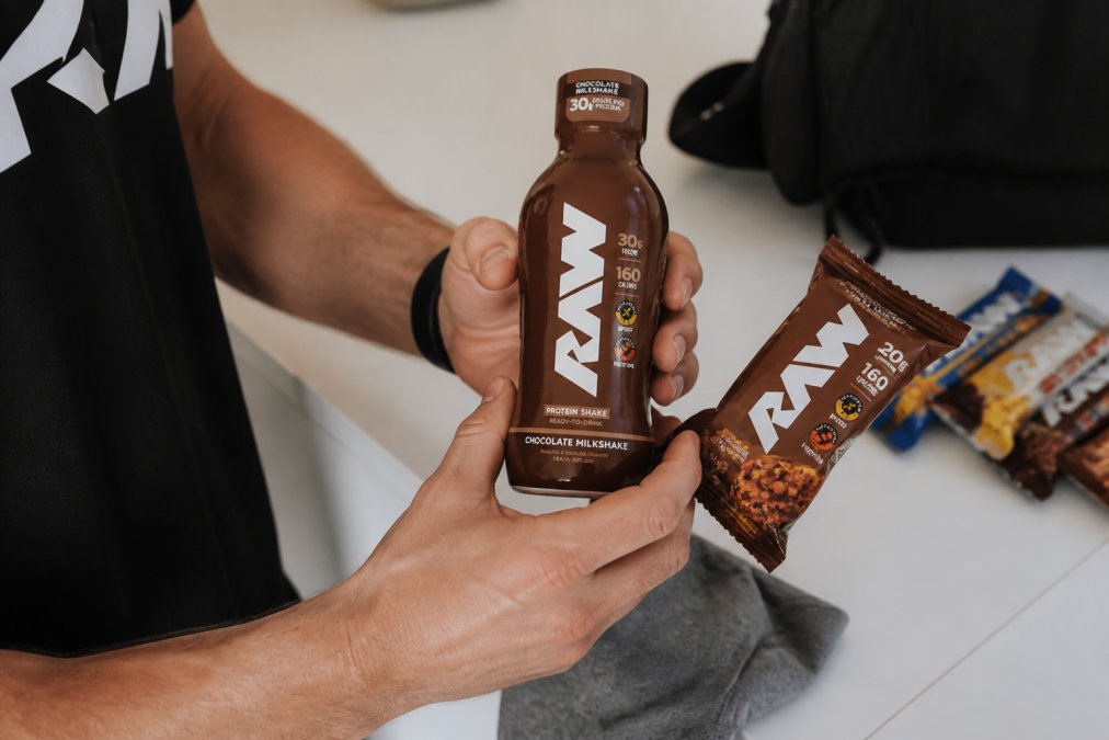 Close-up of hands holding affordable protein bars and ready-to-drink bottles with visible price tags, laid out on white flat surface with natural lighting