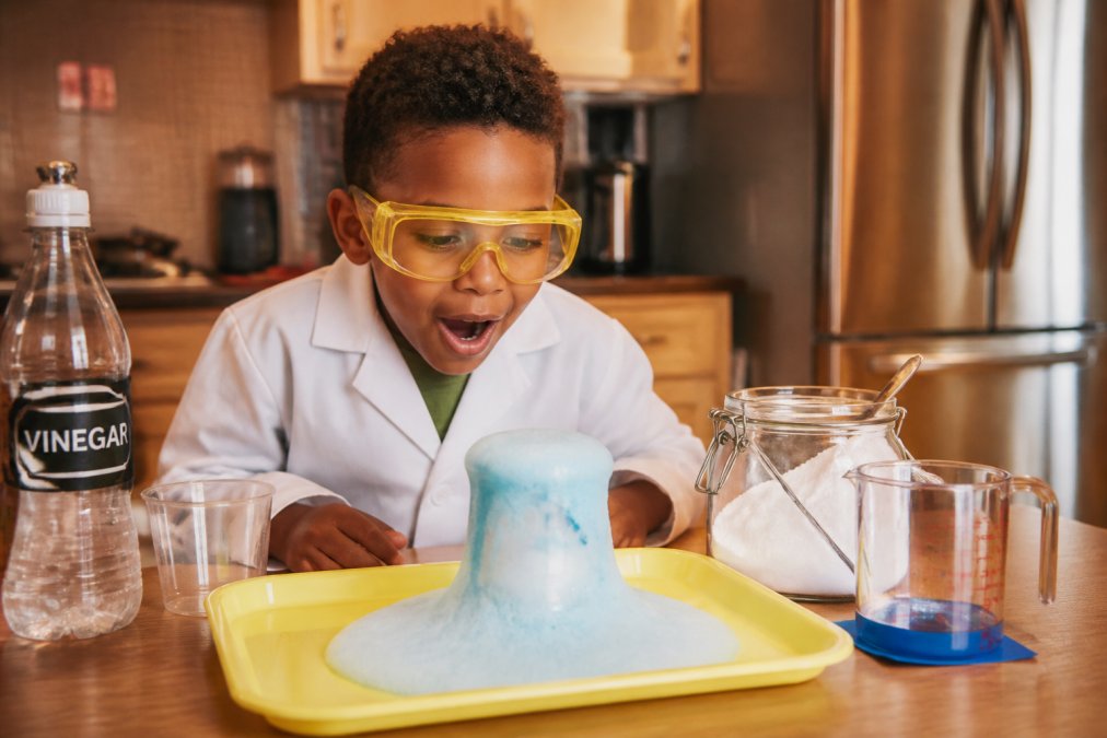 A child at home conducting a simple science experiment at a kitchen table, wearing yellow safety goggles, surrounded by household materials including vinegar, bicarbonate of soda, and measuring cups, looking curious and excited under warm kitchen lighting