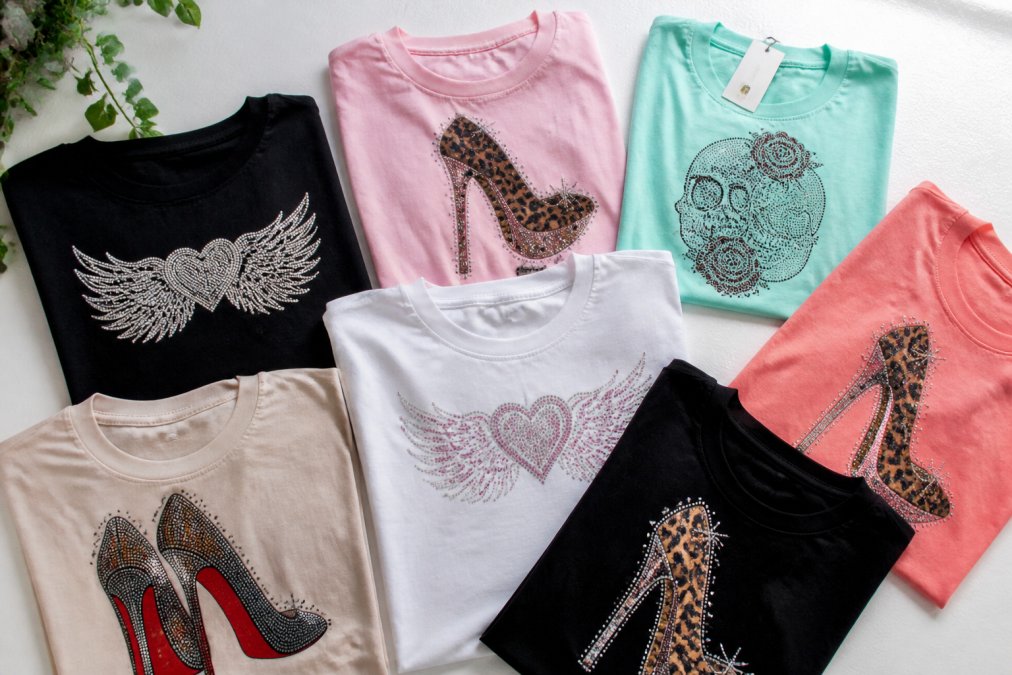 Flat lay of several colorful trendy women's graphic tees and rhinestone-detail tops arranged neatly on a white surface, with a small price tag visible on one item, shot in bright natural light