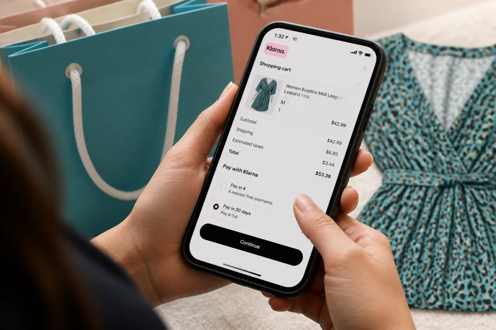 Close-up of hands holding a smartphone displaying a Klarna payment checkout screen with a shopping cart showing dress items, shopping bags visible blurred in background
