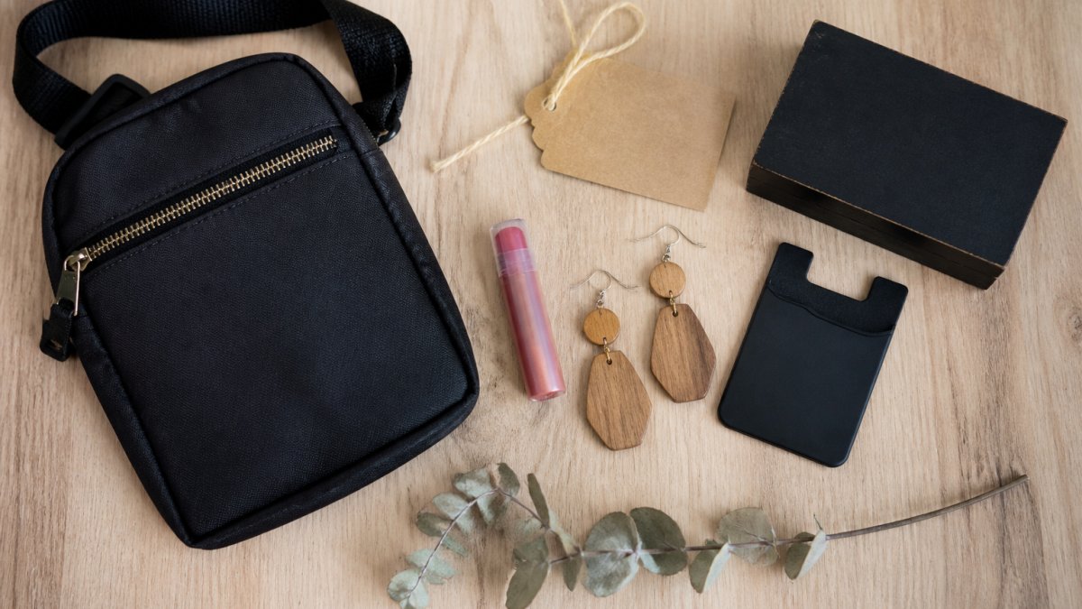 Flat-lay on a light wood table showing a small black crossbody sling bag, a tinted lip balm, wood dangle earrings, a stick-on phone card holder, and a small black wooden box sign, arranged with a handwritten kraft gift tag and a sprig of dried eucalyptus