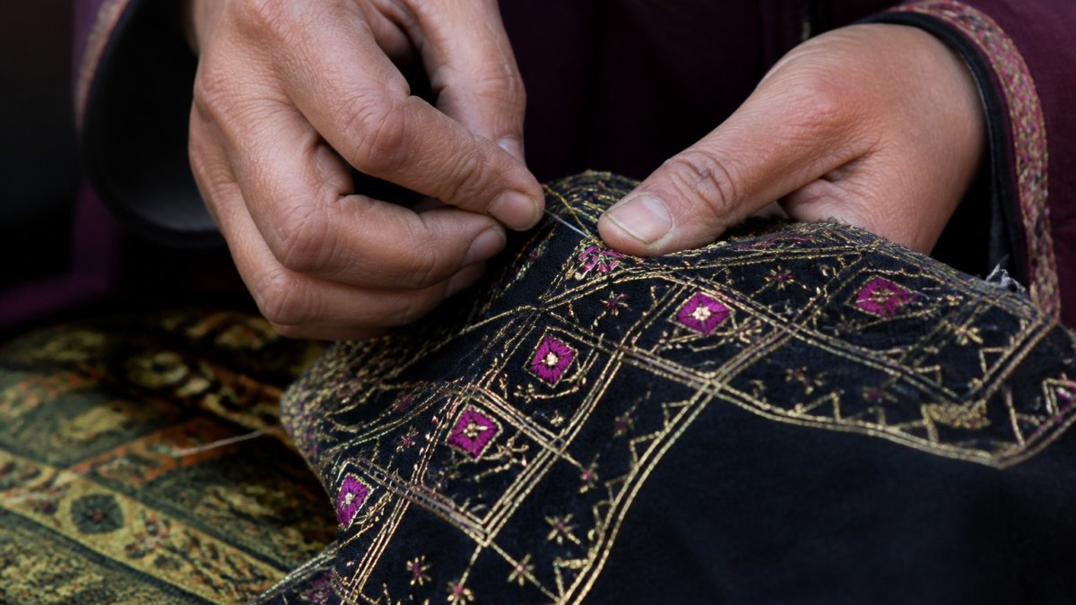 Close-up of hands performing detailed hand embroidery on fabric, showing fine metallic thread being carefully stitched through traditional Afghan textile with intricate geometric patterns