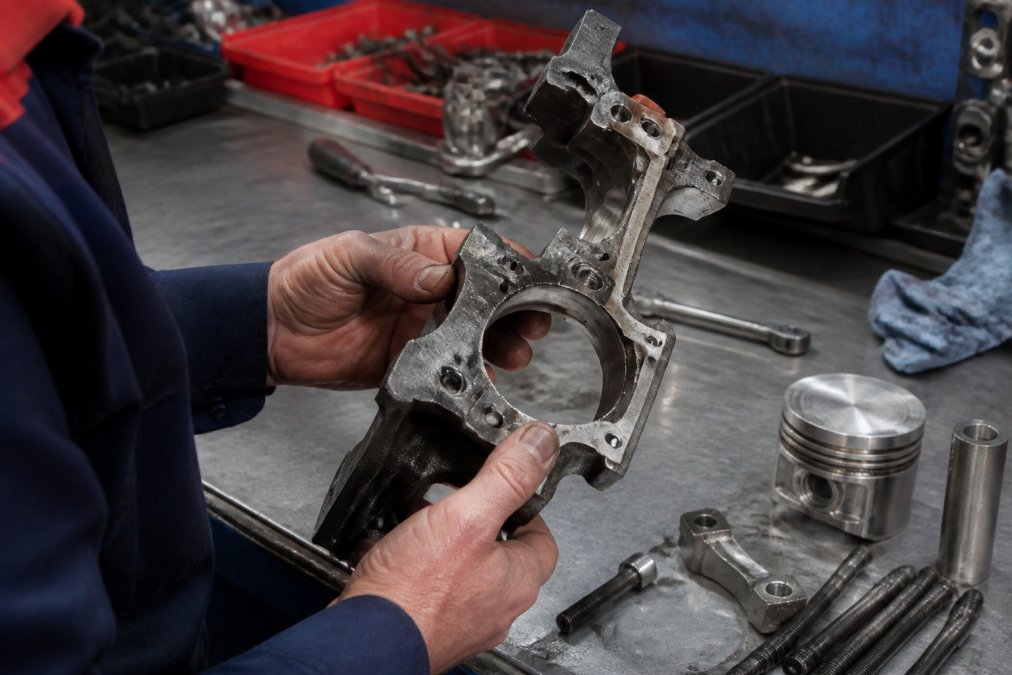 Close-up of a mechanic's hands examining an engine component under bright workshop lighting, checking certification stamps, part numbers, and material quality markings on the part surface