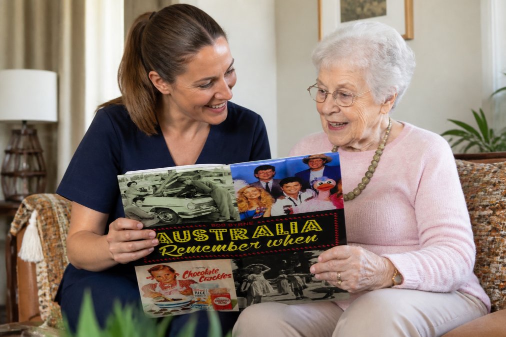 Aged care worker sitting beside an elderly resident in a comfortable lounge room with warm natural lighting, both looking at a nostalgic Australian photo book together, smiling and engaged in conversation