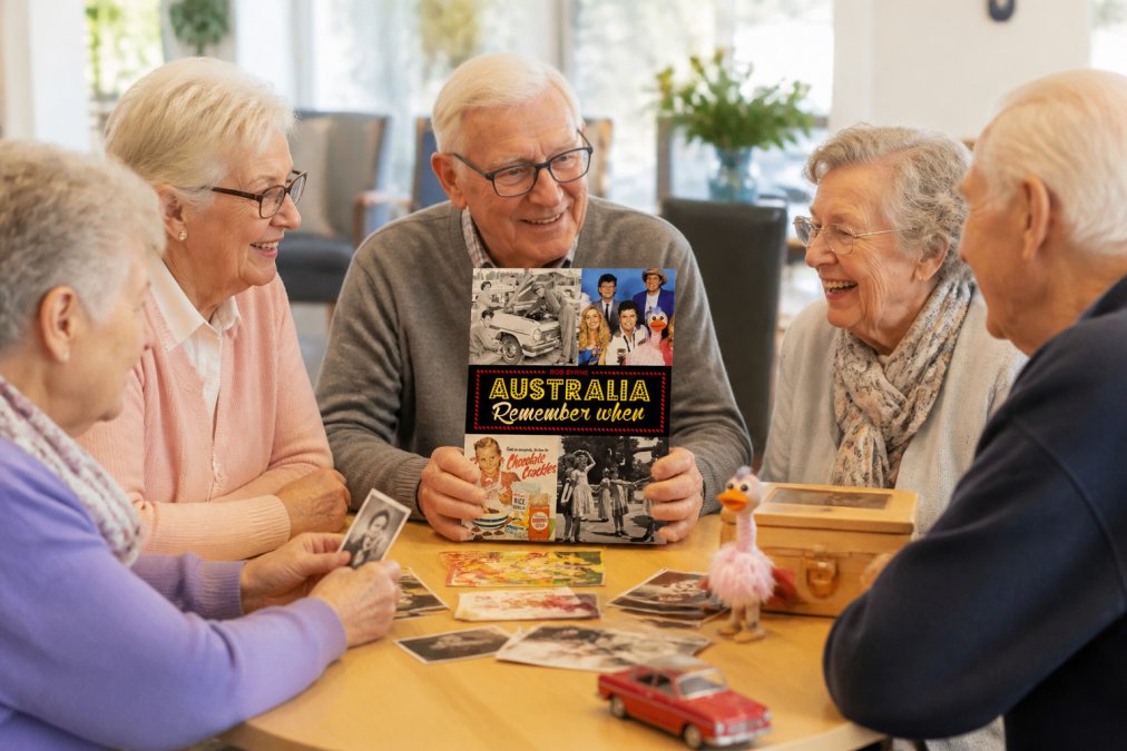 Small group of elderly residents gathered around a table with vintage Australian memorabilia, photographs, and memory prompts, engaged in animated conversation and smiling together