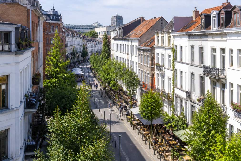 Vue aérienne d'une rue bruxelloise animée avec terrasses, arbres, piétons, et immeubles résidentiels typiques du quartier, lumière du jour naturelle