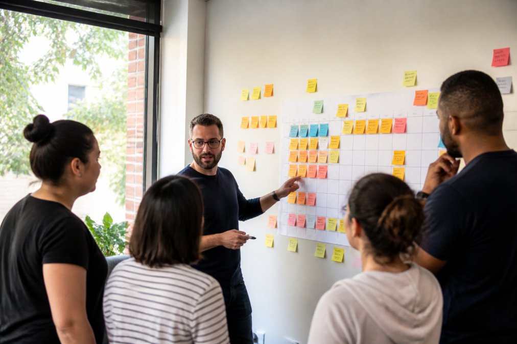 A Scrum Master facilitating a sprint planning session, team gathered around a wall covered in sticky notes and a printed sprint board, bright meeting room