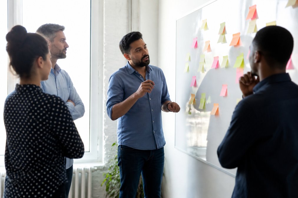 A product owner standing at a whiteboard covered in sticky notes, talking a small cross-functional team through a sprint goal in a bright meeting room