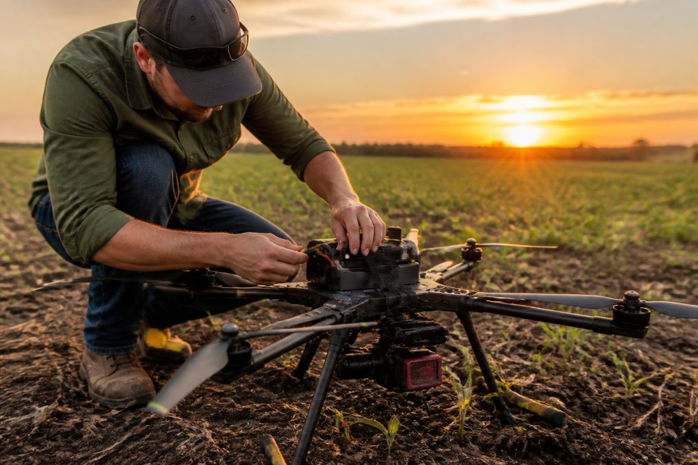 Certified drone pilot conducting pre-flight inspection of multirotor drone equipped with multispectral sensor payload in agricultural field at golden hour, checking battery connections and sensor calibration