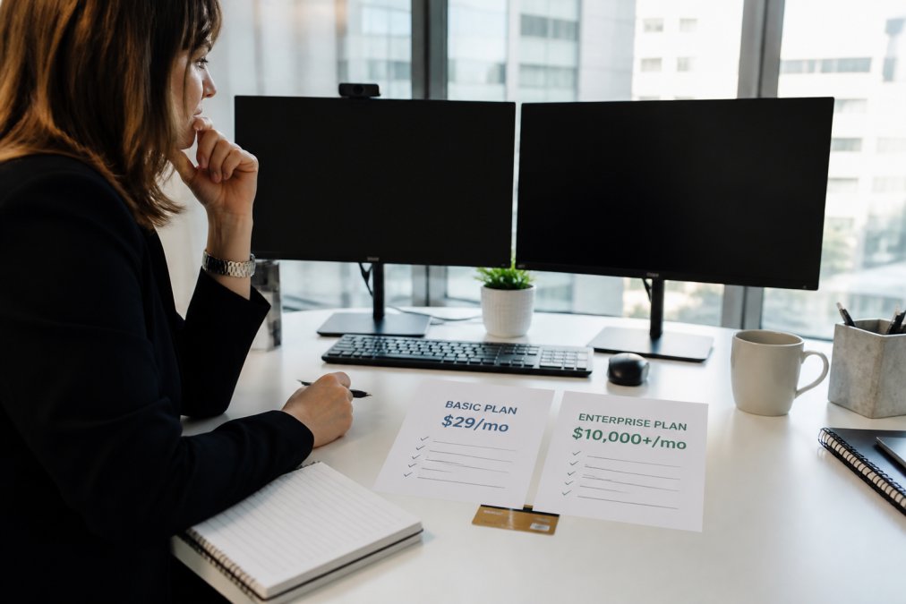 A marketing director at a clean desk comparing software options on two monitors, with a credit card and printed pricing sheets visible in natural office lighting