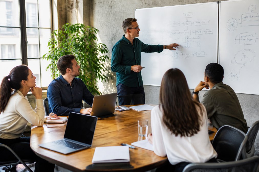 Development team collaborating around a table with laptops and whiteboards showing AI agent architecture diagrams, integration planning, and deployment workflows in a modern tech office with natural lighting