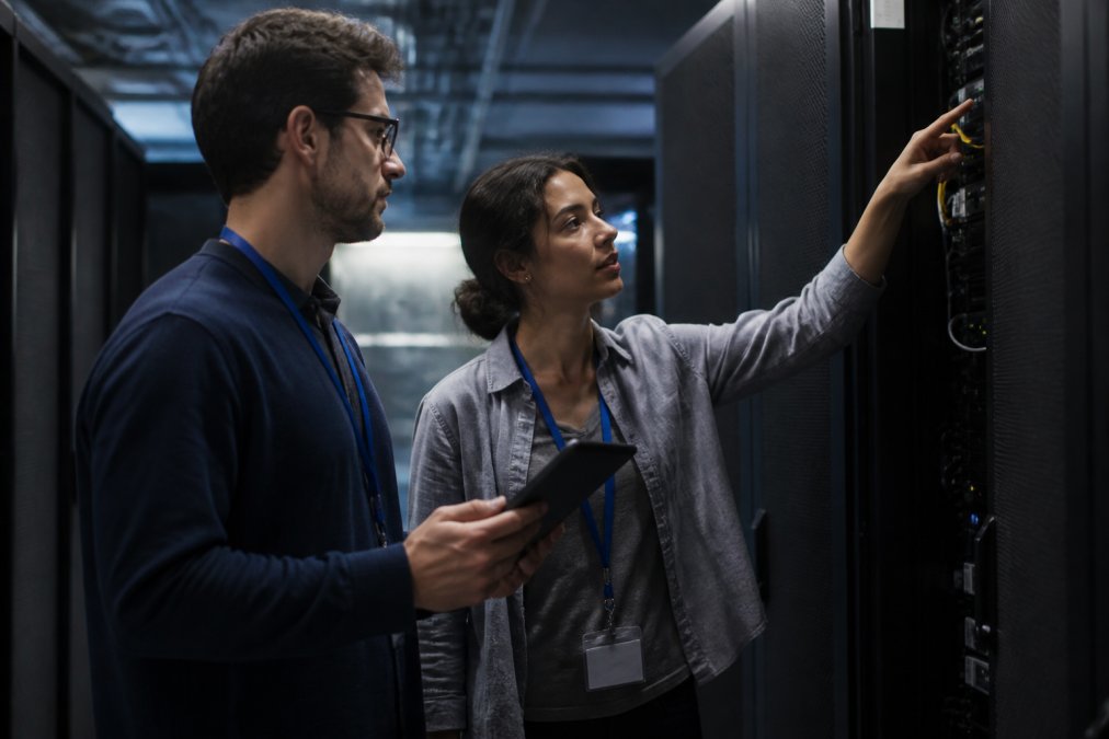 Two security engineers in a dimly lit operations center pointing at a large wall monitor displaying a live network topology map with active agent workflows highlighted in orange, one engineer holding a tablet, blue ambient light from the screens