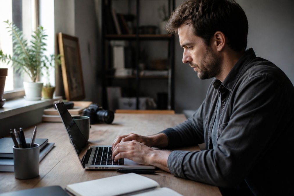 Hands typing detailed design prompt on laptop keyboard with creative workspace visible in background, natural window lighting illuminating the scene
