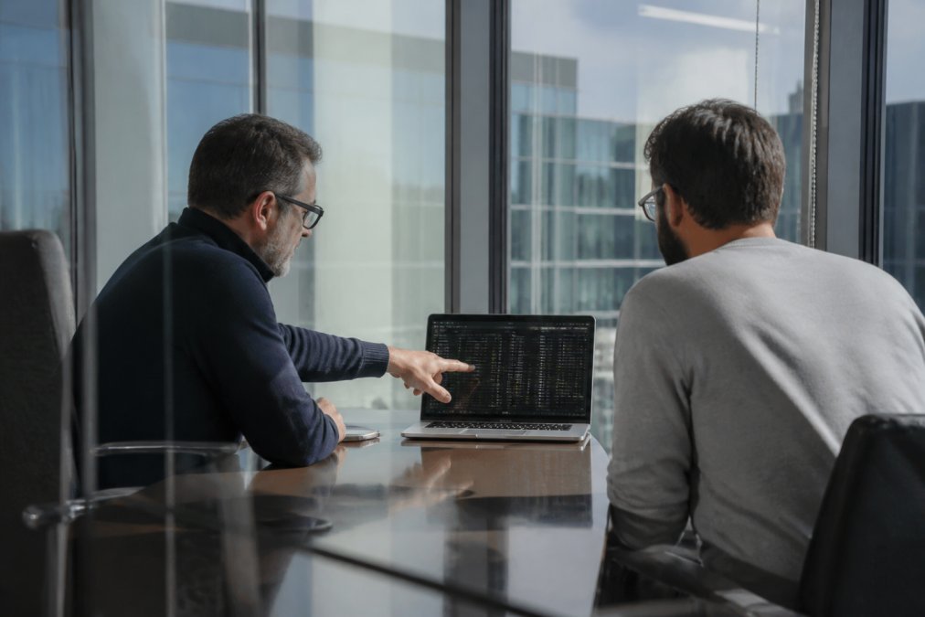 Two security architects in a glass-walled conference room reviewing a laptop screen showing agent execution logs, one pointing at a timeline on the display, cool afternoon light through the windows