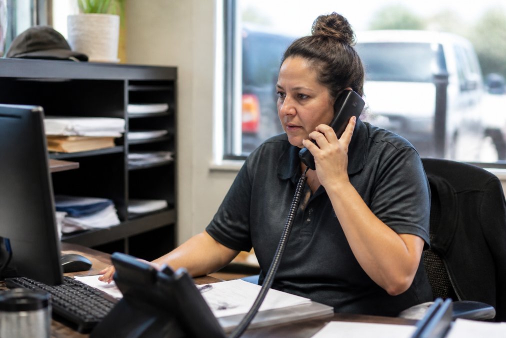 A busy office manager in a small trade business answering a phone call while looking at a scheduling screen on her computer, natural office lighting