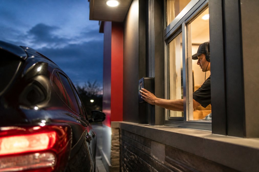 Modern fast food drive-thru window with digital menu board and customer ordering through speaker system at dusk, with crew member visible inside taking orders