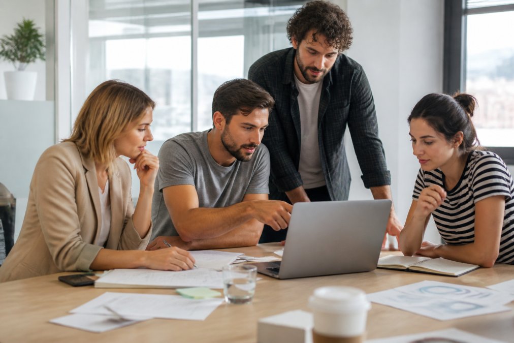 Small marketing team collaborating in modern office with team members pointing at laptop screen displaying campaign analytics while others review notes and strategy documents, bright natural lighting from windows
