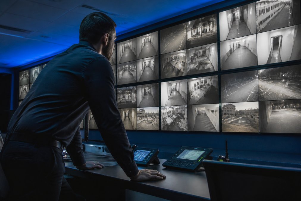 A security professional in a modern control room monitoring multiple live camera feeds on a large display wall, leaning forward with focused attention on the screens, ambient blue lighting from the monitors illuminating the darkened room