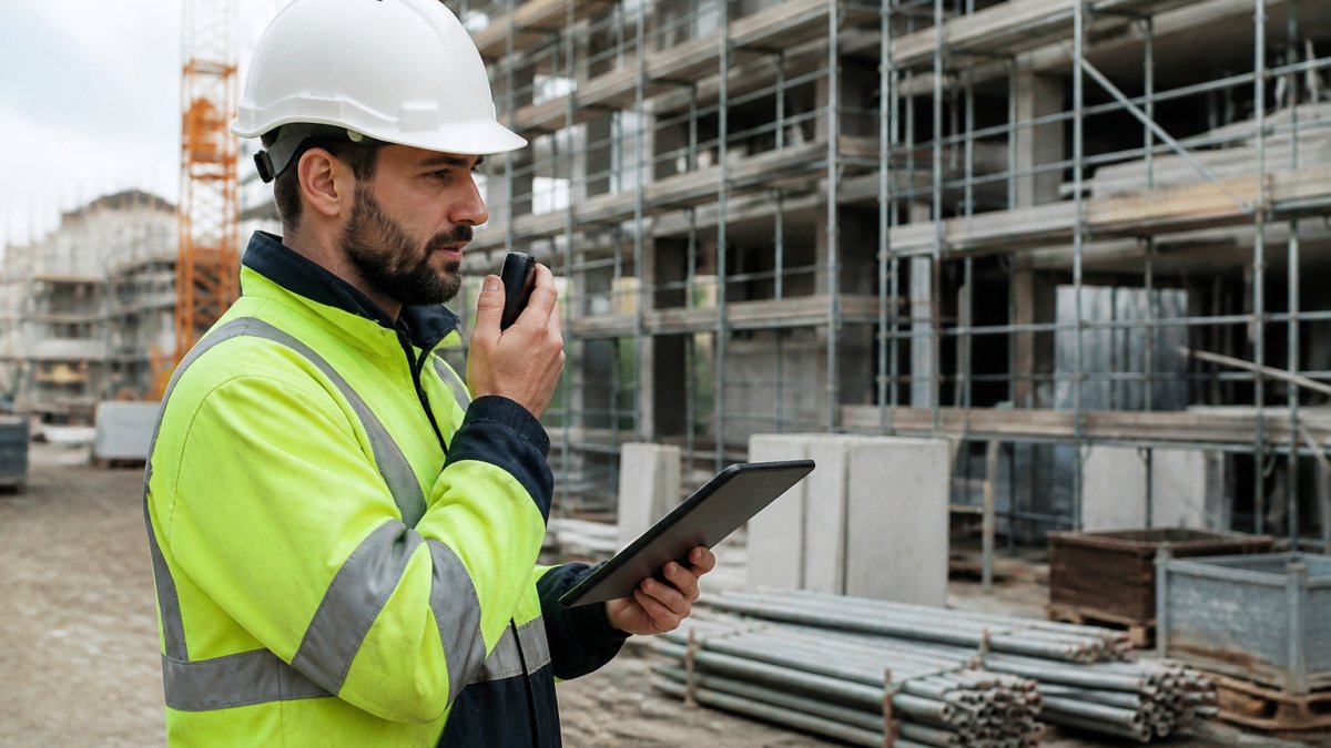 Werfopzichter in veiligheidshelm die op een tablet een spraakmemo inspreekt op een actieve bouwwerf, met bouwmaterialen en steigers op de achtergrond