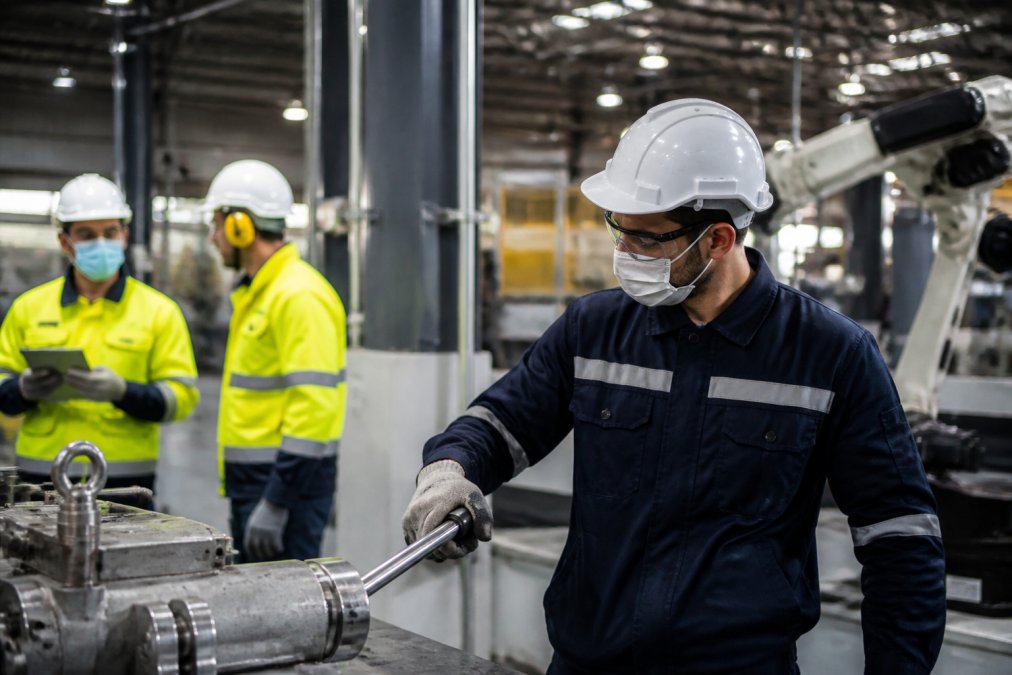 Manufacturing floor with workers wearing hard hats, safety vests, and protective equipment near industrial machinery, with visible safety signage and equipment in a well-lit industrial setting
