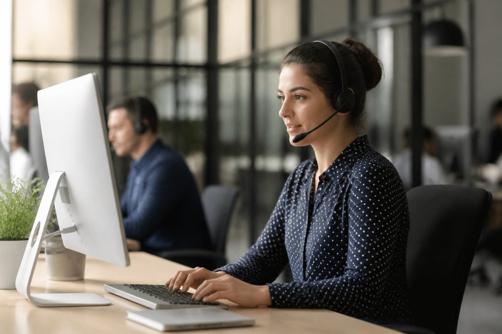 Professional customer service representative at desk with headset, looking at computer screen displaying chat interface with AI-assisted suggestions highlighted, modern office with natural lighting