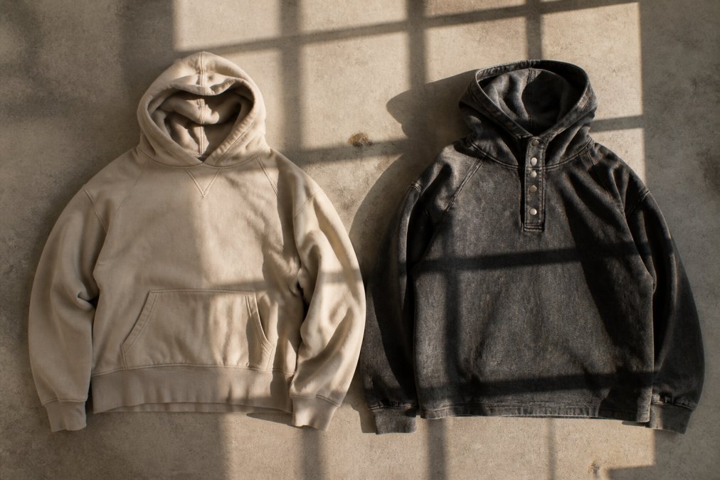 Side-by-side flat lay of two distinct hoodies on a neutral concrete surface: one in muted beige with clean, minimal typography, the other with a bold snow-washed faded graphic and metal snap collar, soft window light casting long shadows