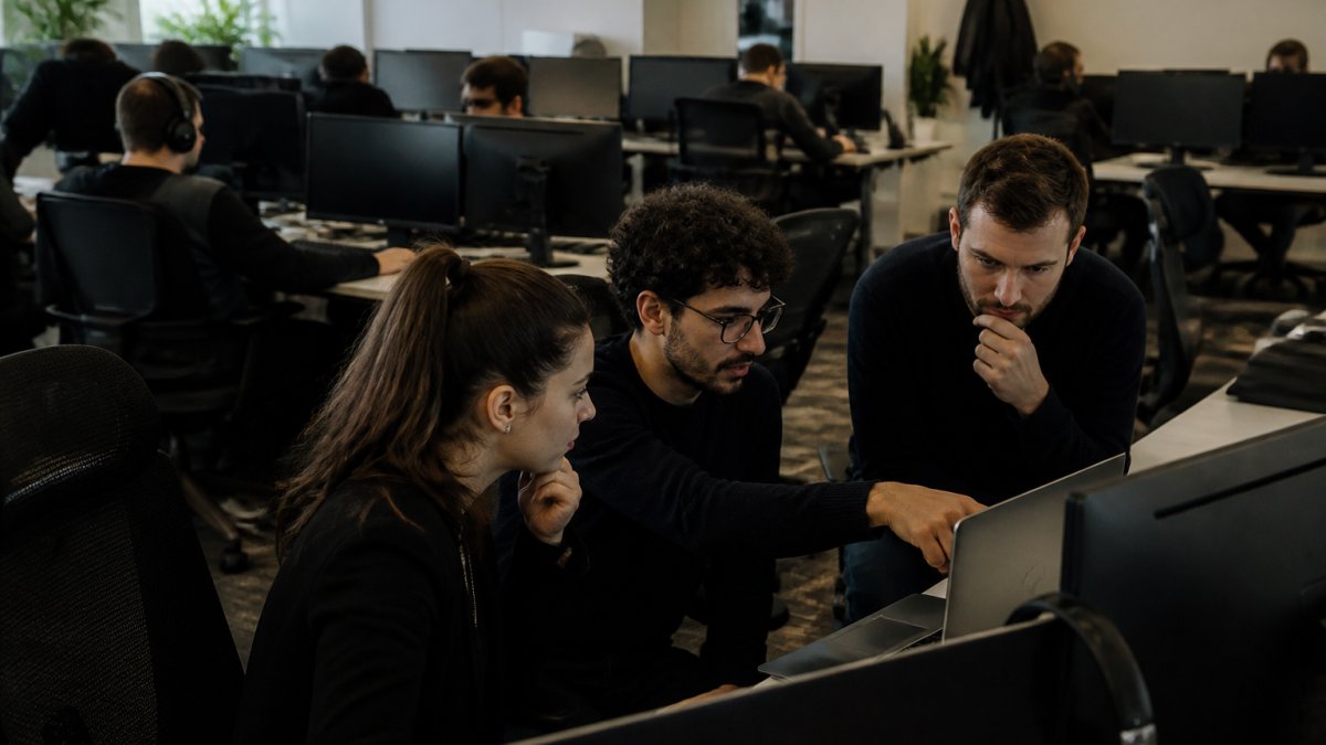 IT operations team monitoring multiple screens in a control room with some staff working at desks and others collaborating on a problem at a workstation, natural lighting from multiple monitors