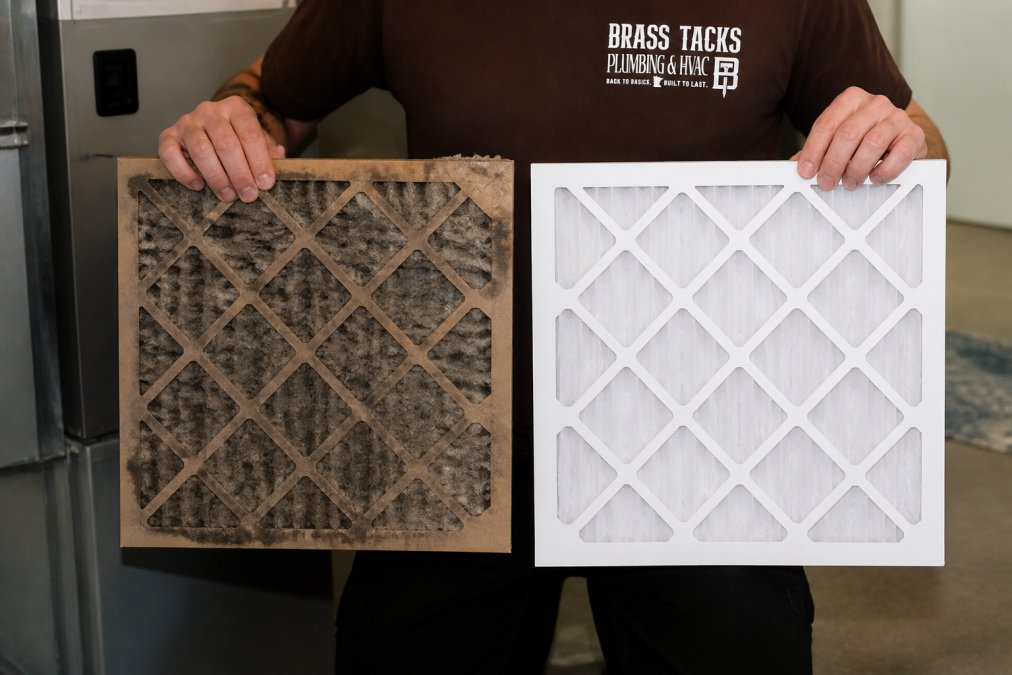 Close-up of a heavily clogged residential HVAC air filter being held up next to a clean white replacement filter, showing the stark contrast in dust and debris buildup under bright indoor lighting