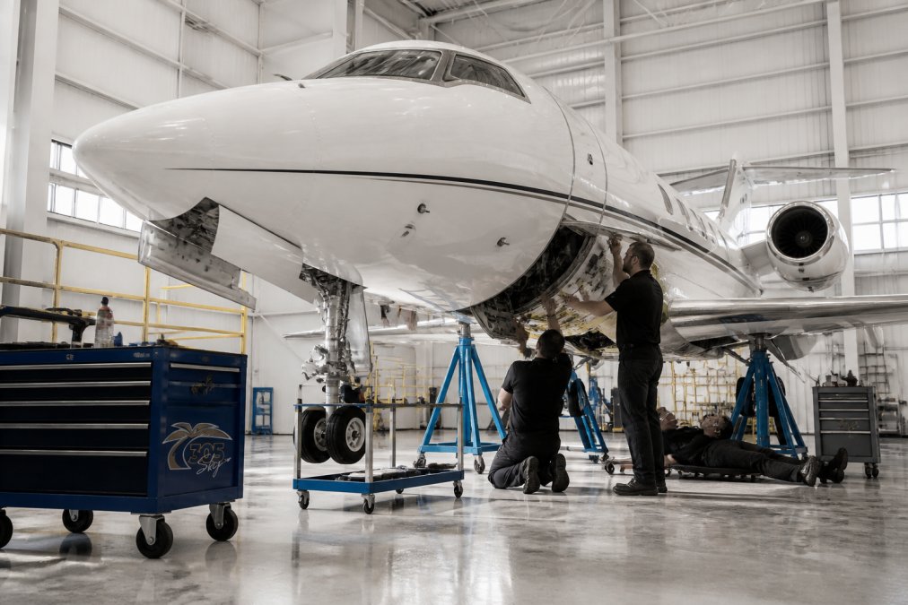 Interior of professional aircraft hangar with ground support equipment, aircraft positioned on maintenance jacks, and maintenance team working on avionics integration and structural inspection under bright overhead lighting
