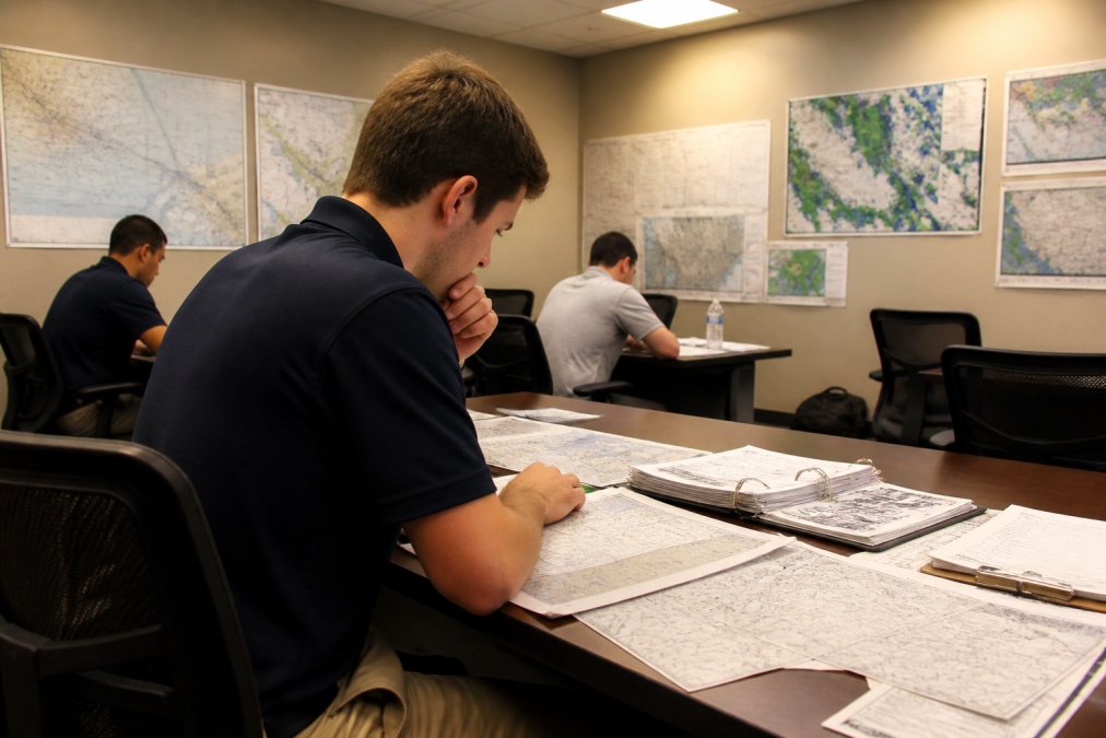 An aircraft dispatcher trainee reviewing a flight plan on a computer screen in a classroom, aviation charts and weather maps pinned to the walls, warm overhead lighting