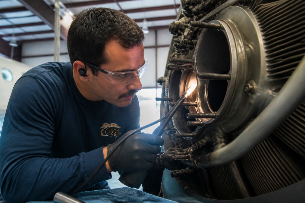 Close-up of an A&P mechanic performing a borescope inspection of an aircraft engine, examining internal cylinder walls and valve seats under bright task lighting in a maintenance hangar