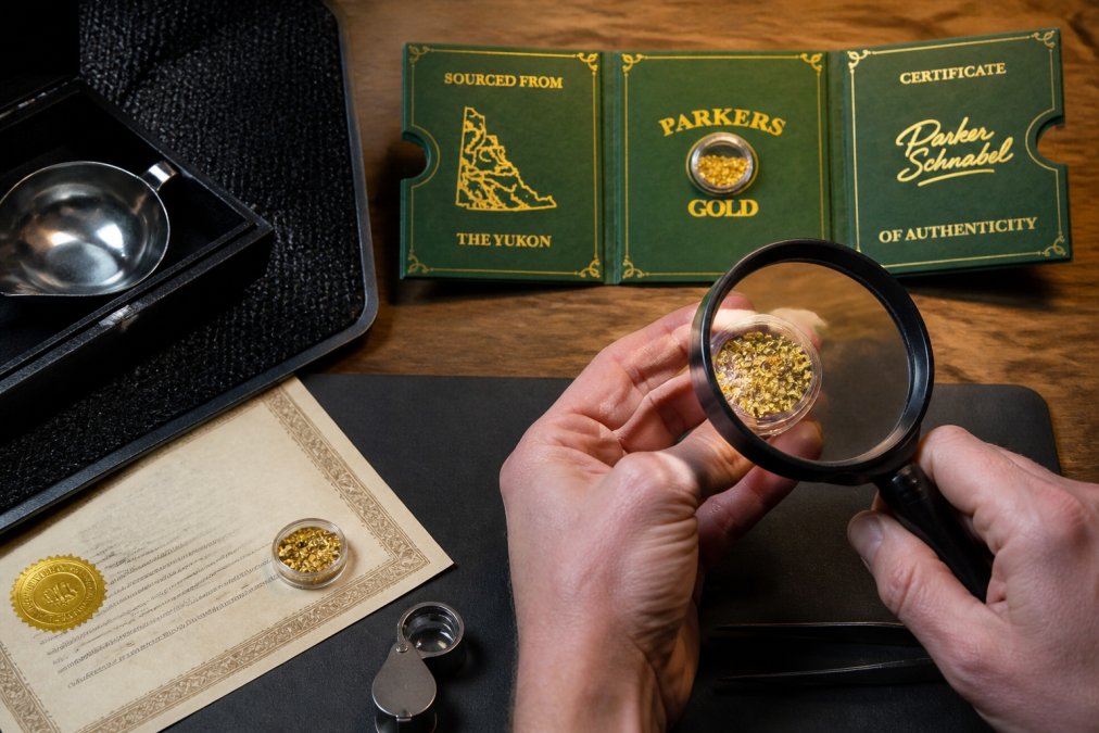 Close-up of hands holding a gold piece under bright magnifying glass and professional lighting, examining surface details and natural texture patterns, with assay certificate visible in background