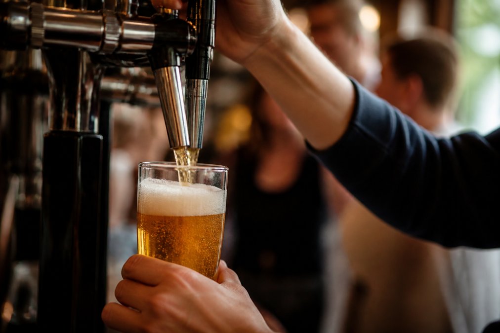 Close-up of an unbranded font pouring a pint of alcohol-free beer into a glass, with golden foam settling at the top, against a warm pub-lighting background