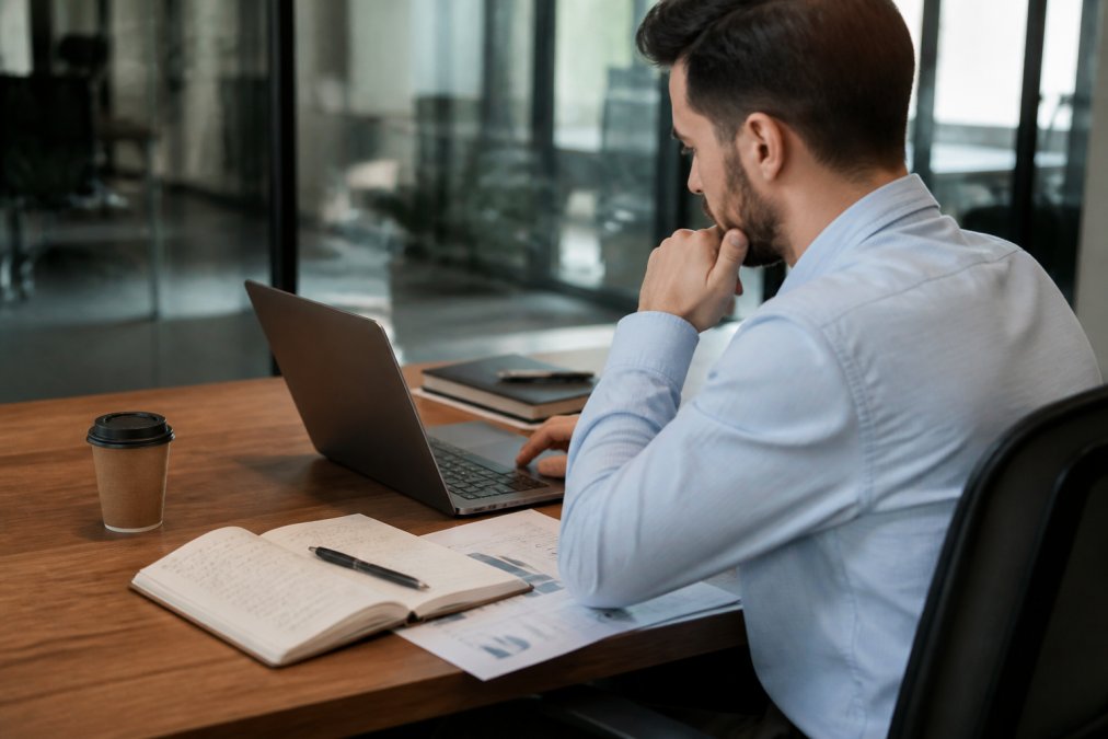 Professional trader analyzing risk metrics and portfolio allocation on a laptop with strategy notes in a notebook nearby, seated at a desk in a modern office environment