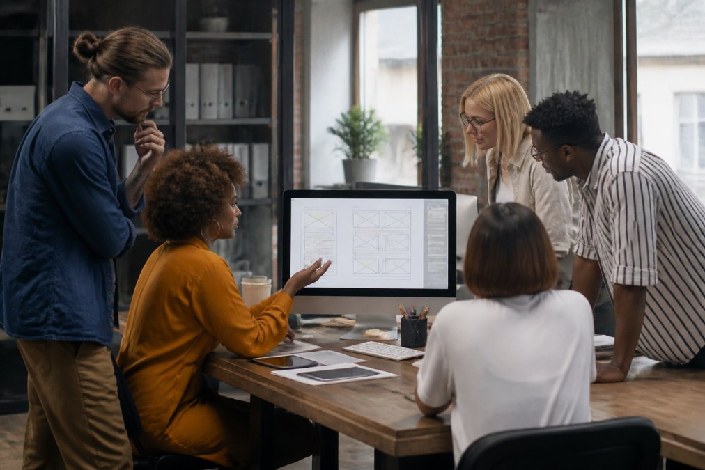 A diverse team of designers and developers collaborating over a large monitor displaying a website wireframe in a bright, modern studio office