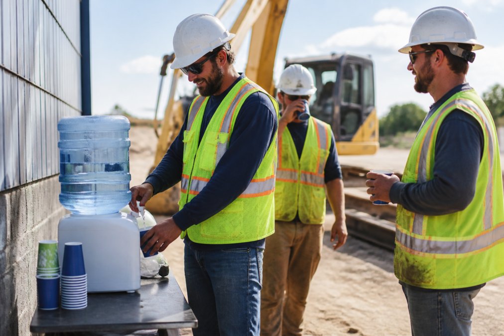 A construction crew taking a water break on a job site, workers in hard hats and safety vests filling cups from a large water dispenser, sunny outdoor setting with heavy equipment in the background