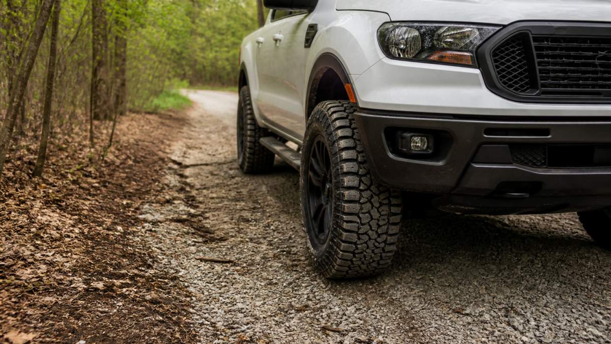 A white pickup truck with aggressive all-terrain tires parked on a gravel road at the edge of a wooded area, with tread blocks visible in the dirt