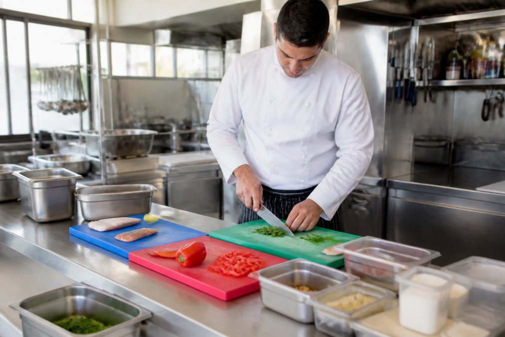 Professional chef in a commercial kitchen preparing food on separate colour-coded cutting boards and prep stations, with clearly labelled allergen containers and dedicated utensils stored separately, natural kitchen lighting
