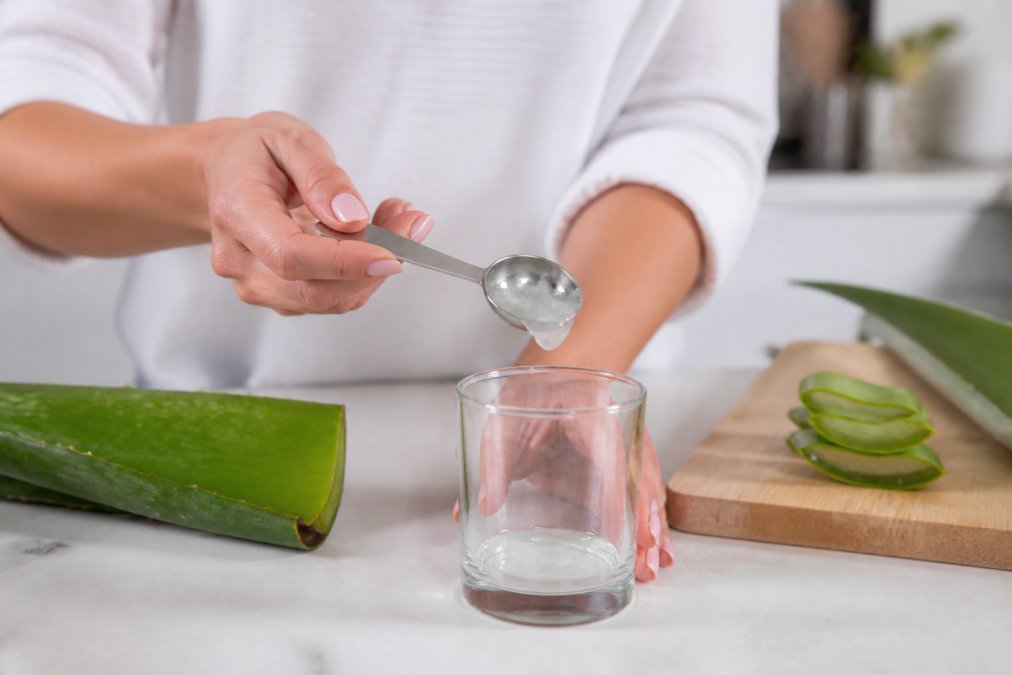 Close-up of hands carefully measuring aloe vera gel into a small clear glass using a measuring spoon, on a clean white kitchen counter with soft natural light