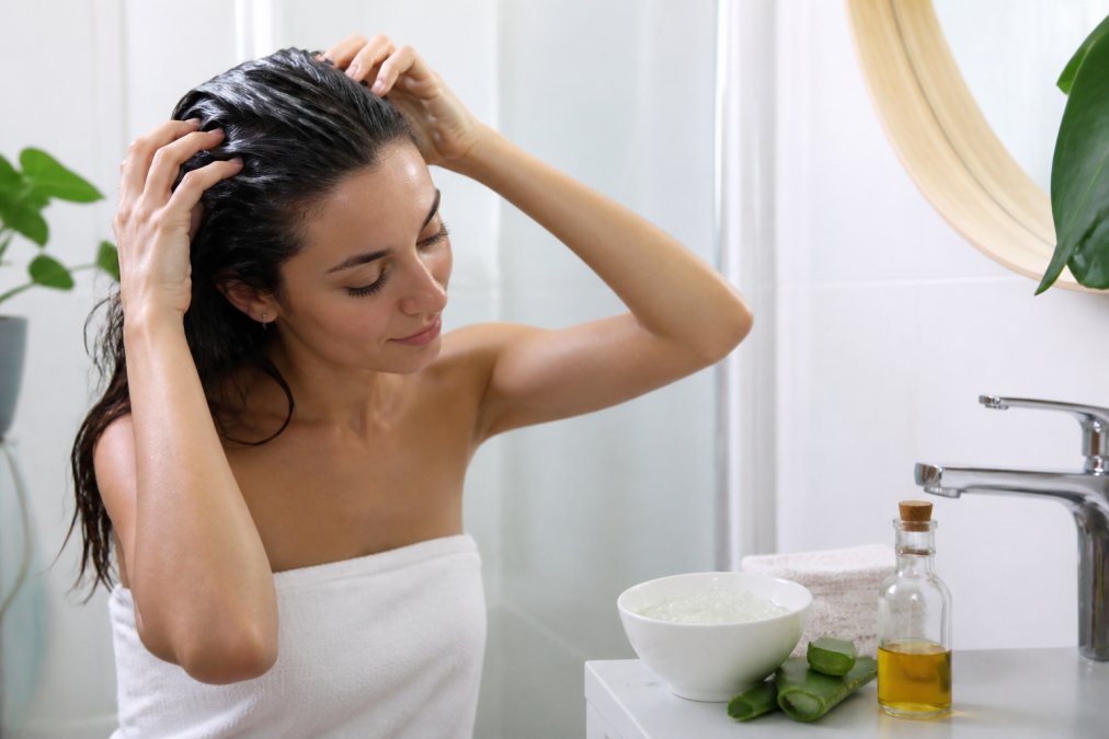 En person står i et lyst badeværelse og påfører en hjemmelavet aloe vera hårkur i håret med hænderne, mens en skål med frisk aloe vera gel og en flaske olie står på vasken