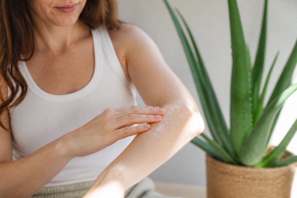 Kvinde påfører klar aloe vera gel på tør hud på underarmen i naturligt lys, med grøn aloe vera plante synlig i baggrunden