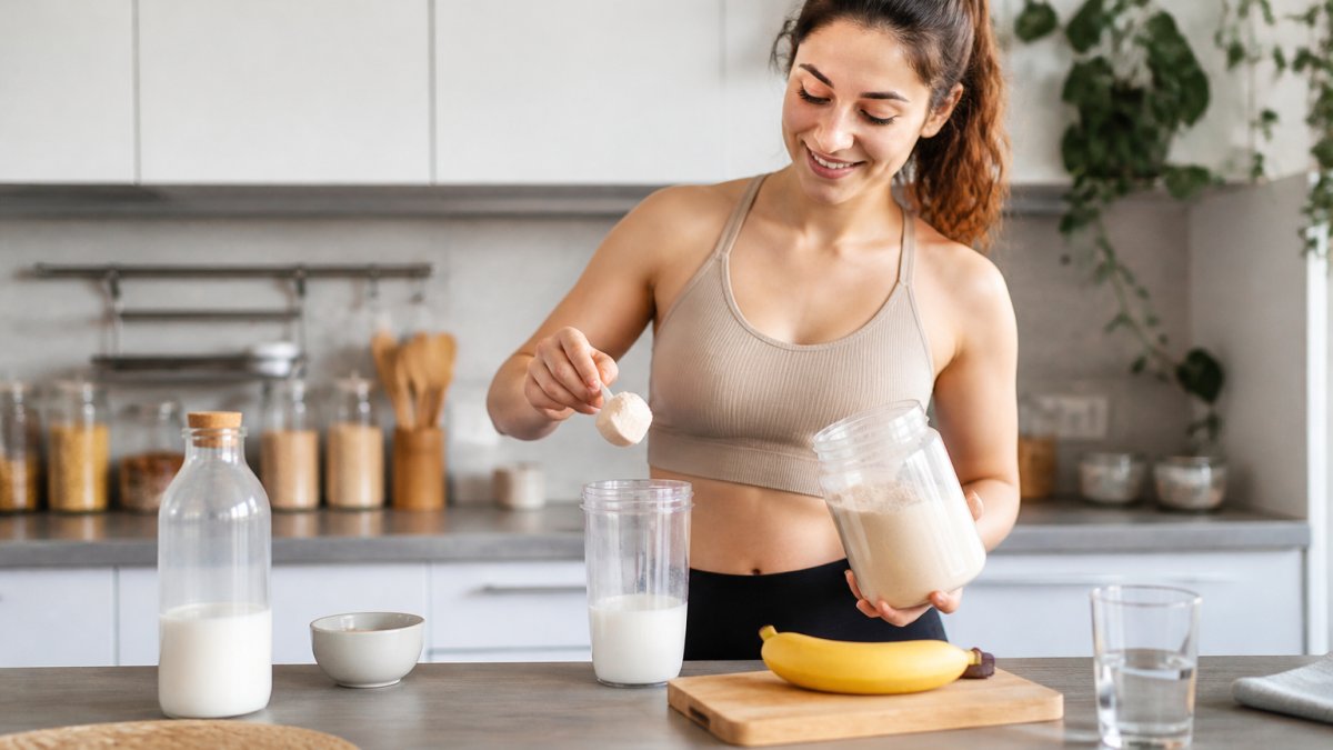 Person mixing protein powder with almond milk and a banana in a bright kitchen