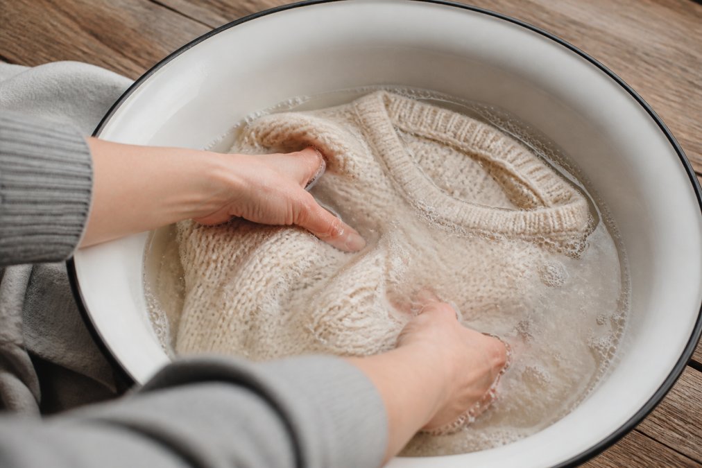 Close-up of hands gently submerging a cream-colored alpaca sweater in a basin of cool water with mild soap, showing proper hand-washing technique with soft, controlled movements