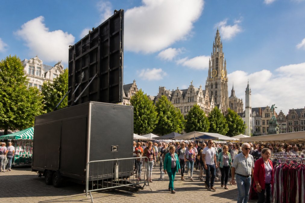 Mobiele LED-aanhangwagen met groot scherm op een druk openluchtmarkt in Antwerpen, met publiek, marktkramen en stadsachtergrond zichtbaar in zonnig weer