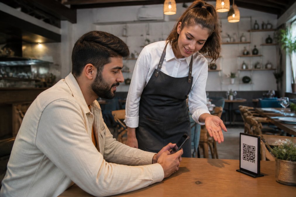 Camarero en un restaurante moderno mostrando a un cliente cómo acceder al menú digital mediante código QR desde su smartphone, con pantalla clara visible en la mesa de madera