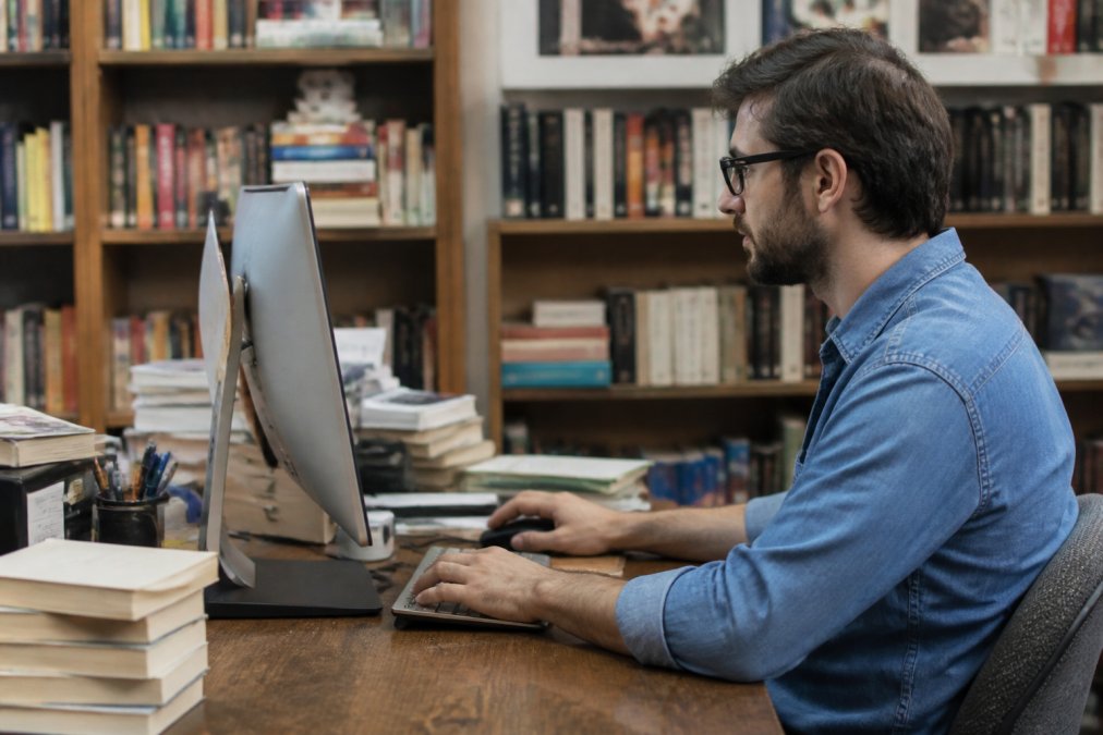 Libraio che lavora al computer in negozio con scaffali di libri sullo sfondo, consultando il gestionale cloud su schermo con focus naturale su display e mani sulla tastiera