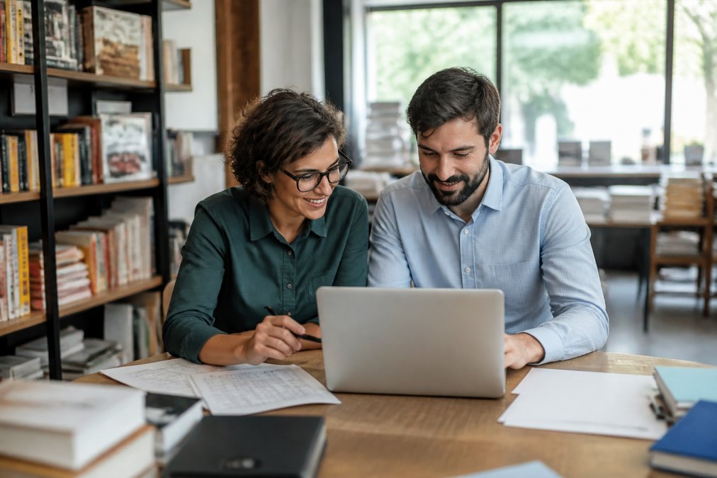 Due persone che lavorano insieme su un laptop durante una sessione di onboarding, con documenti e appunti sulla scrivania, in ufficio luminoso con finestre naturali